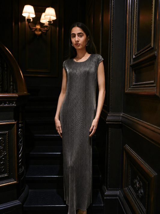 A woman stands on an indoor staircase wearing a sleeveless, floor-length textured dress in a dark charcoal gray color. The background features dark wood paneling and wall sconces.