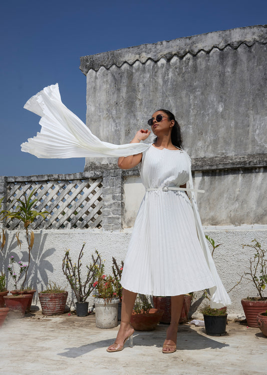 A woman wearing a white pleated dress with cloak sleeves and a belt, standing on a rooftop surrounded by potted plants.