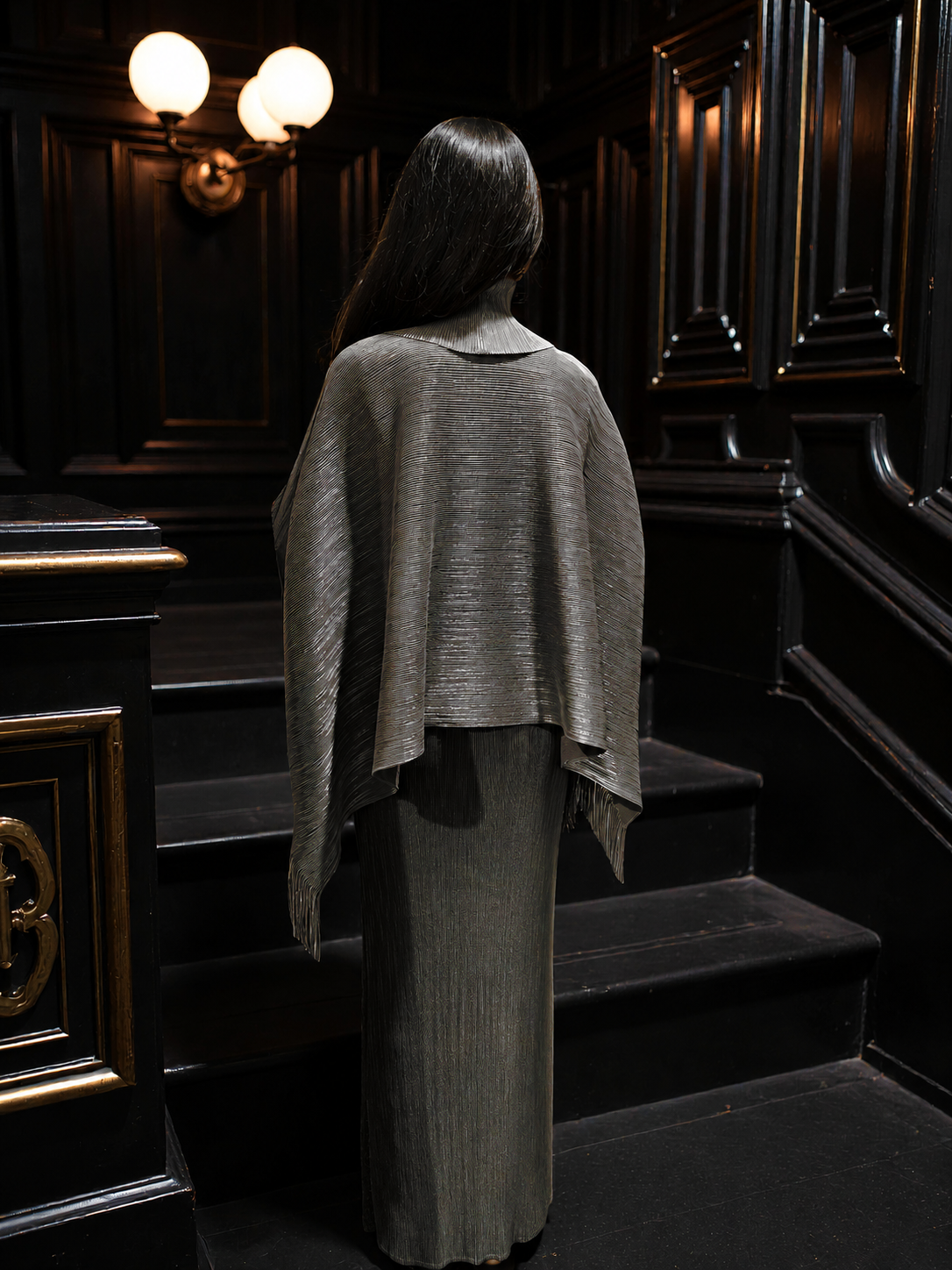 A woman viewed from behind wearing a textured, draped gray shawl and matching pleated skirt, standing on dark wooden stairs in a dimly lit interior.