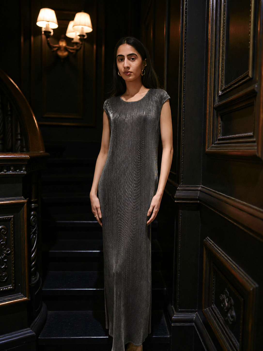 A woman stands on an indoor staircase wearing a sleeveless, floor-length textured dress in a dark charcoal gray color. The background features dark wood paneling and wall sconces.