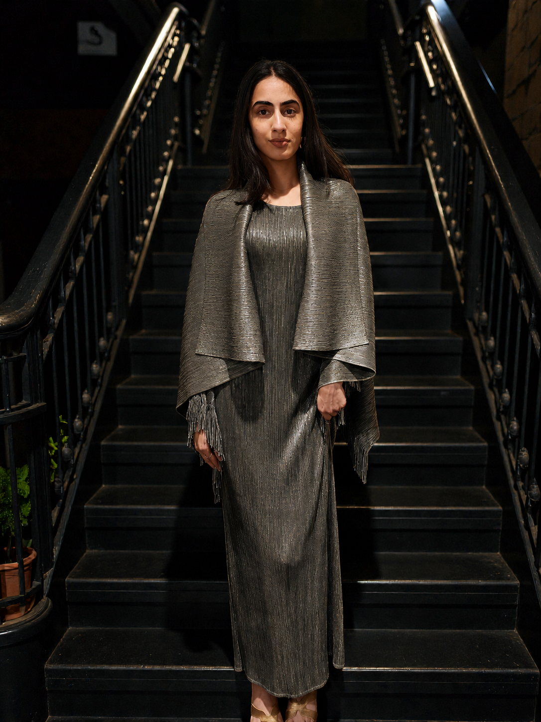 A woman stands on a dark staircase wearing a metallic silver pleated maxi dress with a matching draped cape-style shawl featuring fringe details.