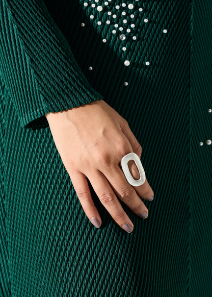 A close-up of a hand wearing a chunky, minimalist white ring with an open oval shape, set against a dark green textured knit dress with rhinestone embellishments.