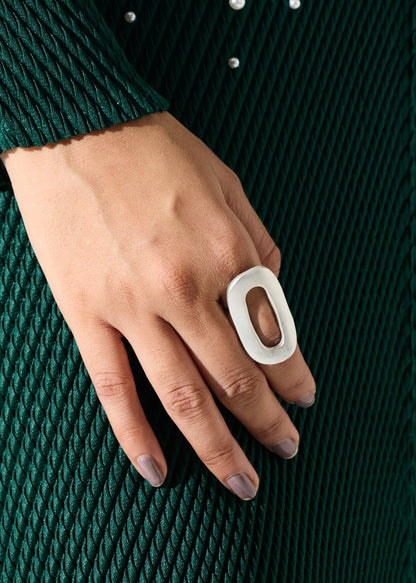 A close-up of a hand wearing a chunky, oversized white ring with a hollow oval center, set against a dark green textured knit sweater.