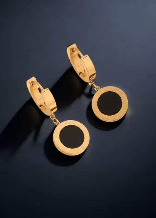 A pair of gold hoop earrings with circular drop pendants featuring a black center, displayed on a dark navy blue reflective surface.