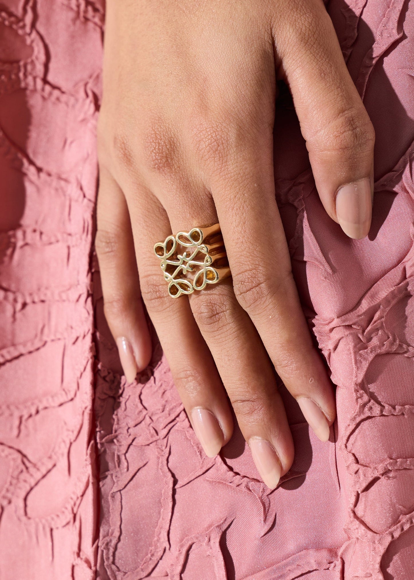 A close-up of a hand wearing a gold ring with three interlocking openwork circular motifs, resting against a textured pink fabric background.