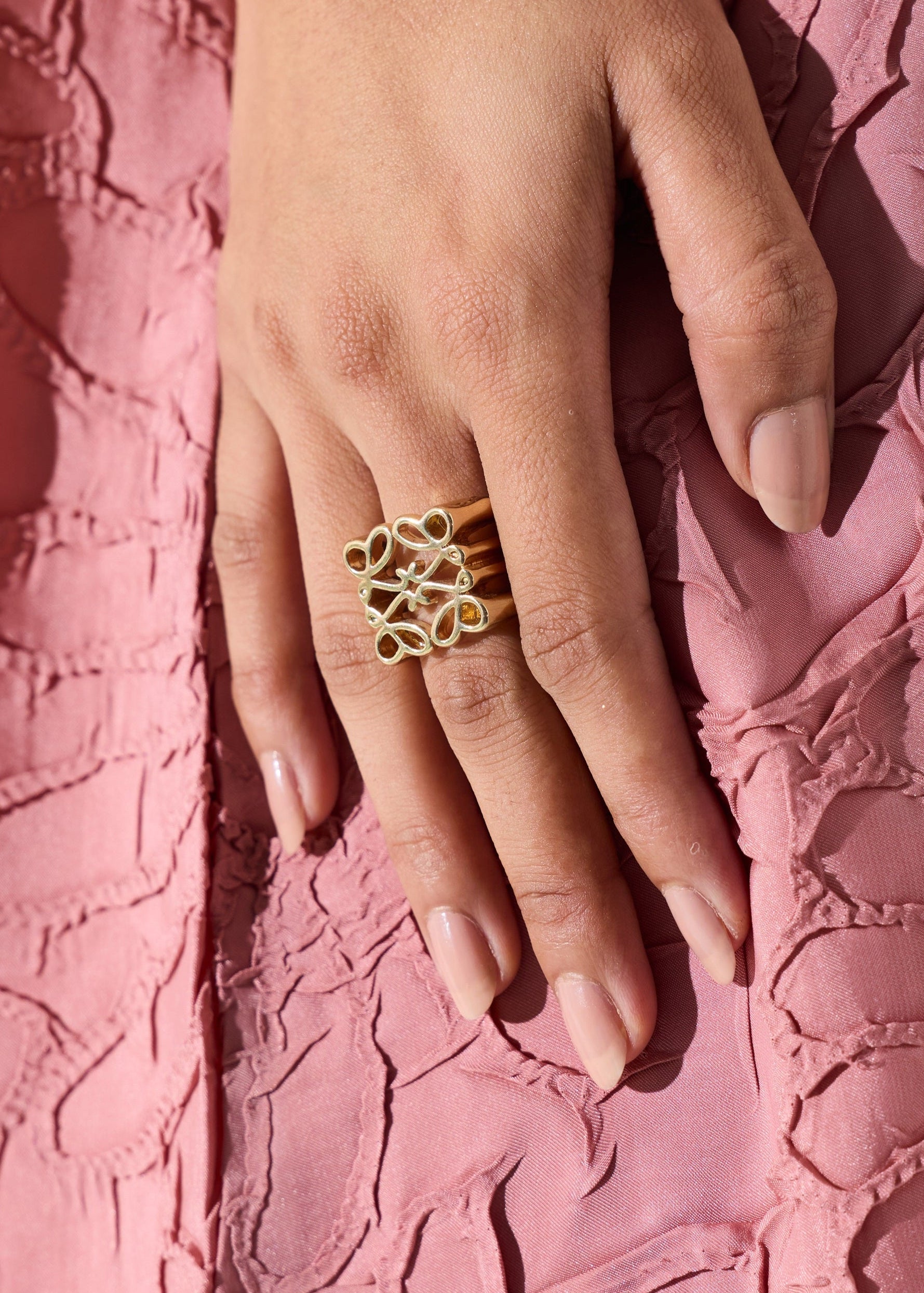 A close-up of a hand wearing a gold ring with three interlocking openwork circular motifs, resting against a textured pink fabric background.