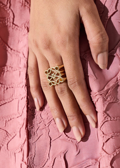 A close-up of a hand wearing a gold ring with three interlocking openwork circular motifs, resting against a textured pink fabric background.