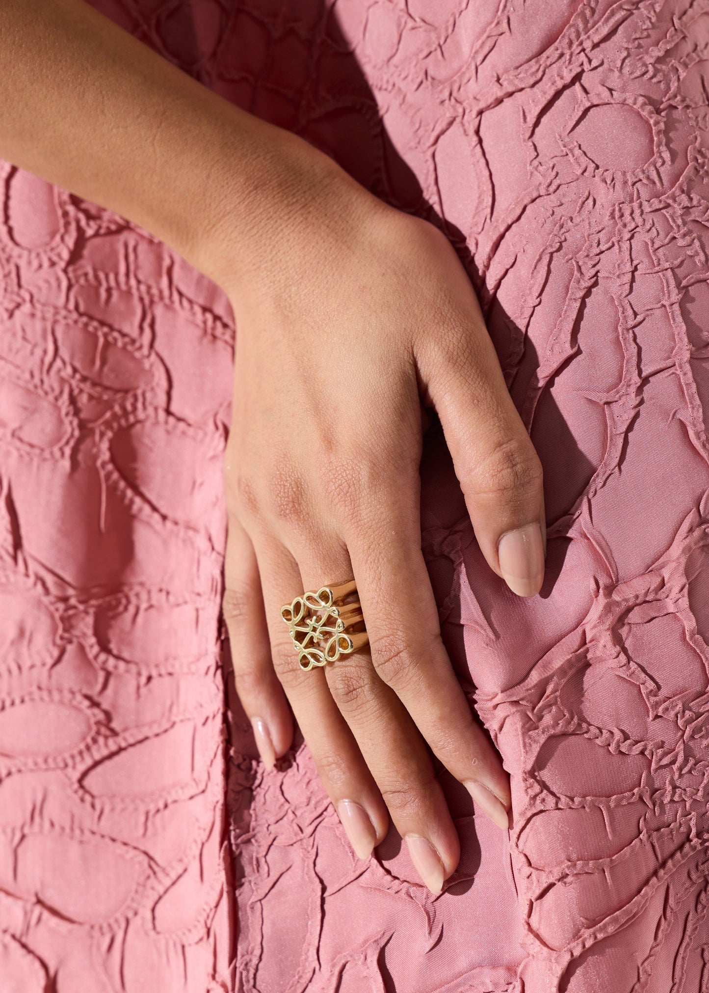 A close-up of a hand wearing a gold ring with a floral or butterfly-like openwork design, resting on a textured pink dress.
