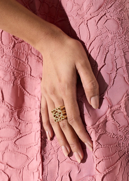 A close-up of a hand wearing a gold ring with a floral or butterfly-like openwork design, resting on a textured pink dress.