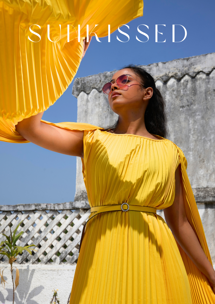 Woman in a yellow dress with sunglasses against a blue sky and stone wall background, featuring the brand 'Sunkissed'.