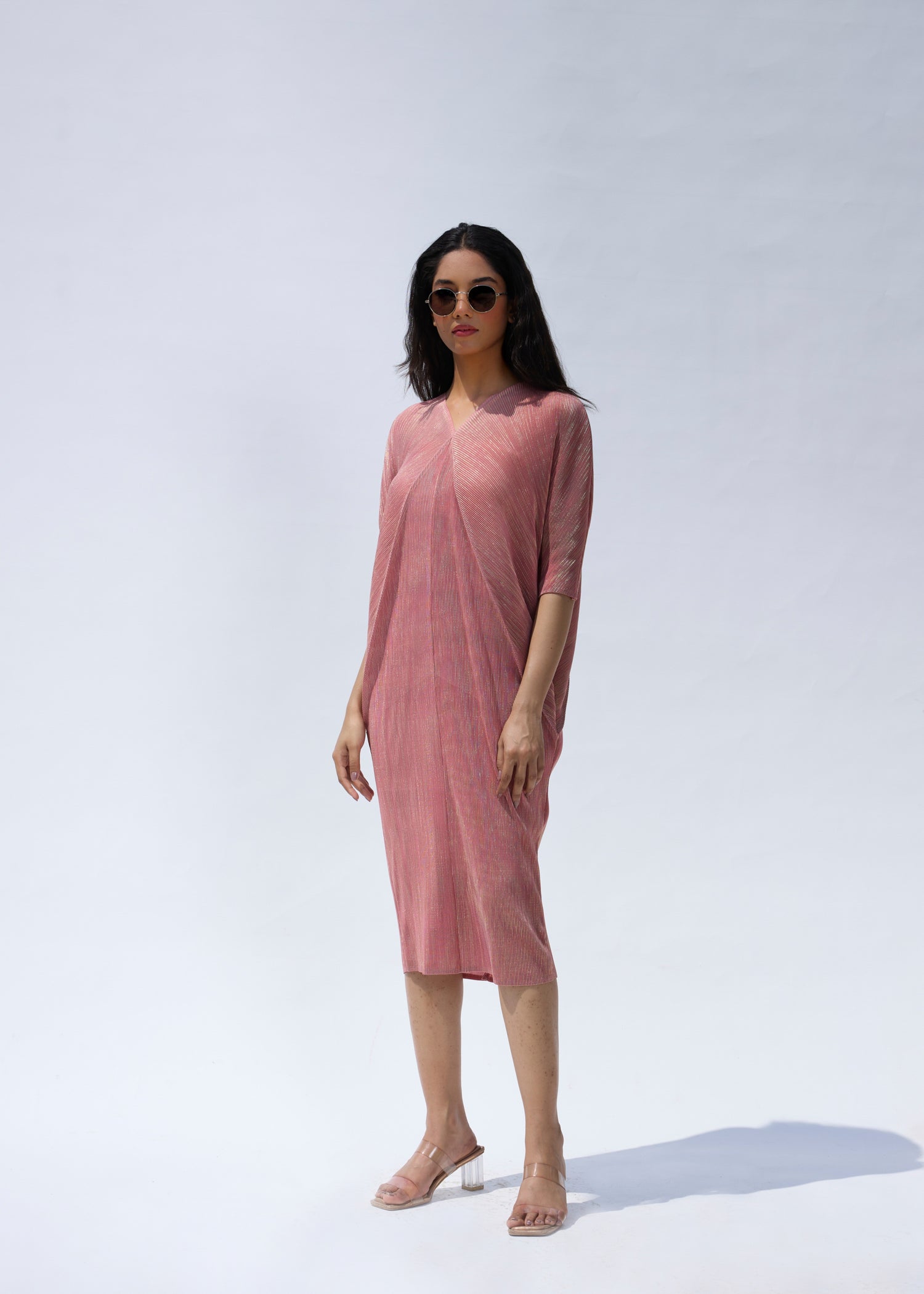 A woman standing in a serene pose wearing a knee-length pink pleated dress with a V-neckline and three-quarter sleeves.