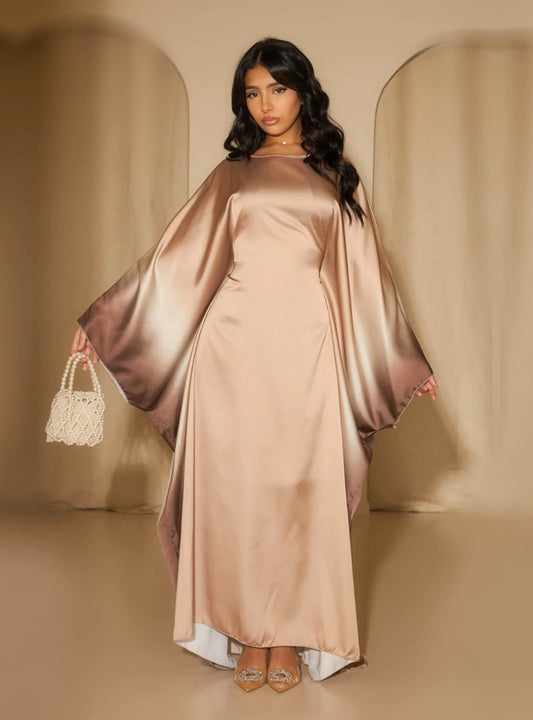 Woman in a long, flowing dress with a cape holding a small handbag against a beige background